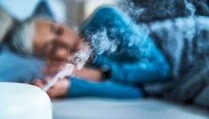 A woman sleeps next to a humidifier shooting steam into the bed room.