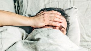 A man waking up and putting his hand to his forehead and grimacing.