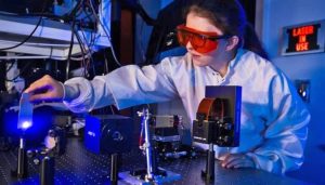 A researcher wearing a white lab coat and red safety goggles uses a laser device in a lab.